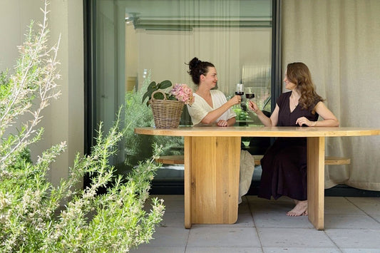 Two women sitting at the ETAUDORE Agata table, raising glasses with Moon & June in a relaxed terrace setting.
